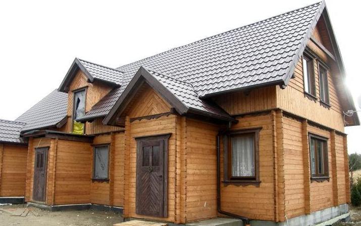 Surowy stan zewnętrzny domu z drewnianych bali i elementami konstrukcji szkieletowej, z widoczną metalową dachówką i niezabezpieczonym oknem na poddaszu.